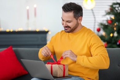 Photo of Celebrating Christmas online with exchanged by mail presents. Happy man opening gift box during video call on laptop at home