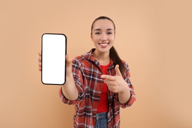 Young woman showing smartphone in hand and pointing at it on light brown background