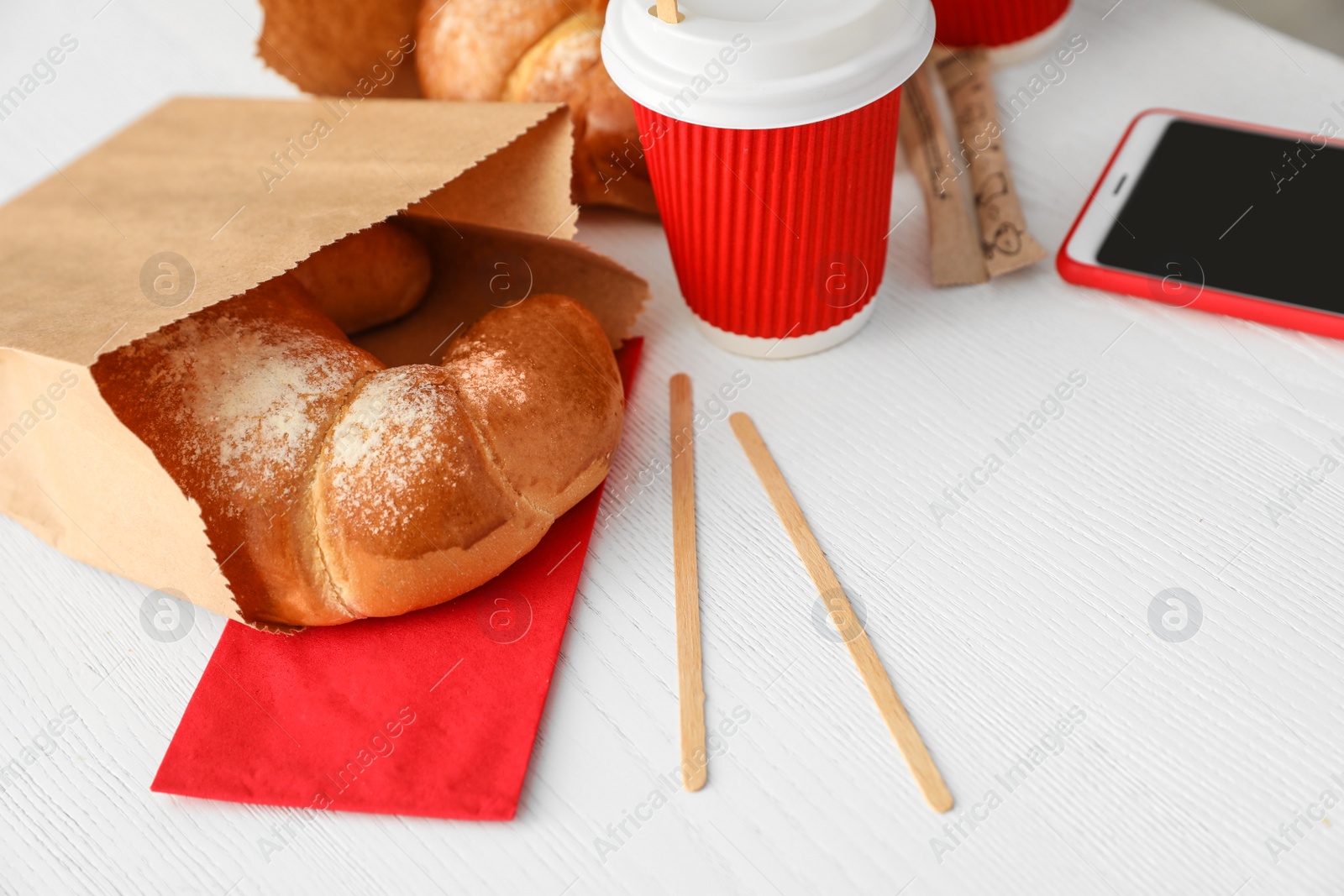 Photo of Cup of coffee, bun in paper bag and smartphone on wooden table