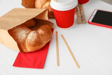 Cup of coffee, bun in paper bag and smartphone on wooden table