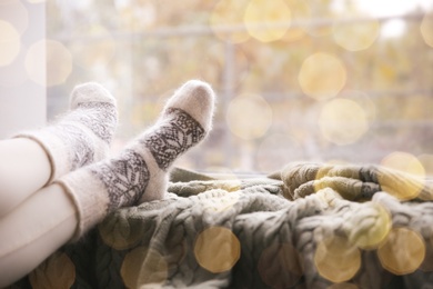 Image of Woman wearing warm socks on knitted plaid near window, closeup. Space for text