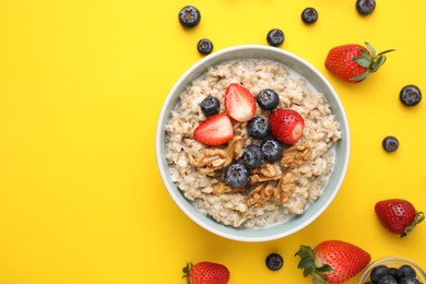 Photo of Tasty oatmeal with strawberries, blueberries and walnuts in bowl surrounded by ingredients on yellow background, flat lay. Space for text