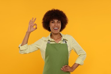 Photo of Happy young woman in apron showing ok gesture on orange background