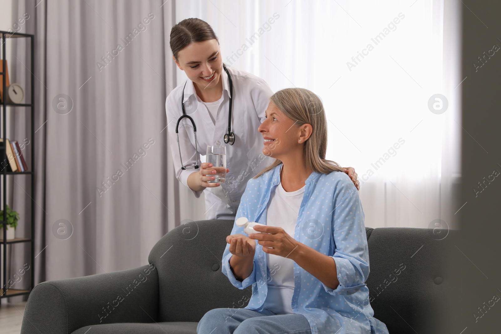 Photo of Young healthcare worker giving glass of water to senior woman with pills indoors