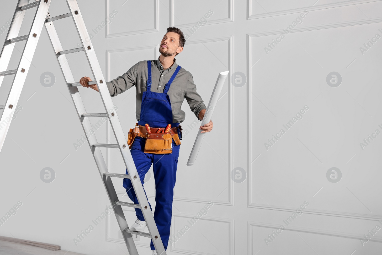 Photo of Electrician in uniform with ceiling lamp on metal ladder indoors