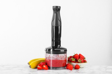 Hand blender with mixture of ingredients and fresh fruits on white marble table