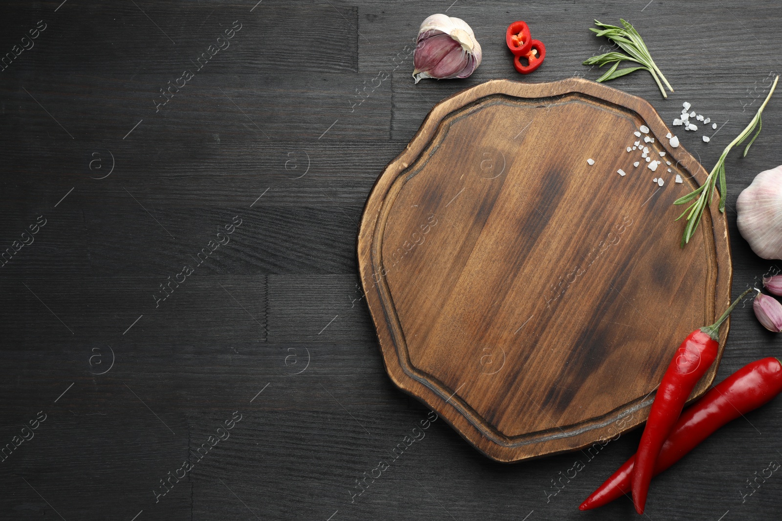 Photo of Cutting board, garlic, rosemary and chili peppers on black wooden table, flat lay. Space for text