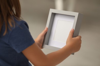 Photo of Little girl holding empty photo frame on blurred background, closeup