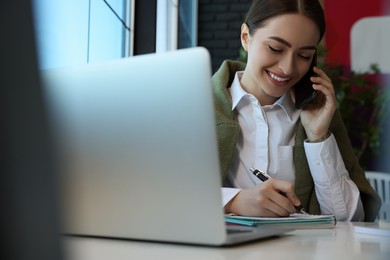 Photo of Young female student with laptop talking on phone while studying at table in cafe