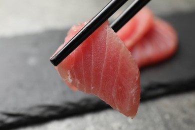 Photo of Holding tasty sashimi (piece of fresh raw tuna) with chopsticks against blurred background, closeup