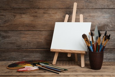 Photo of Easel with blank canvas, palette and brushes on wooden table