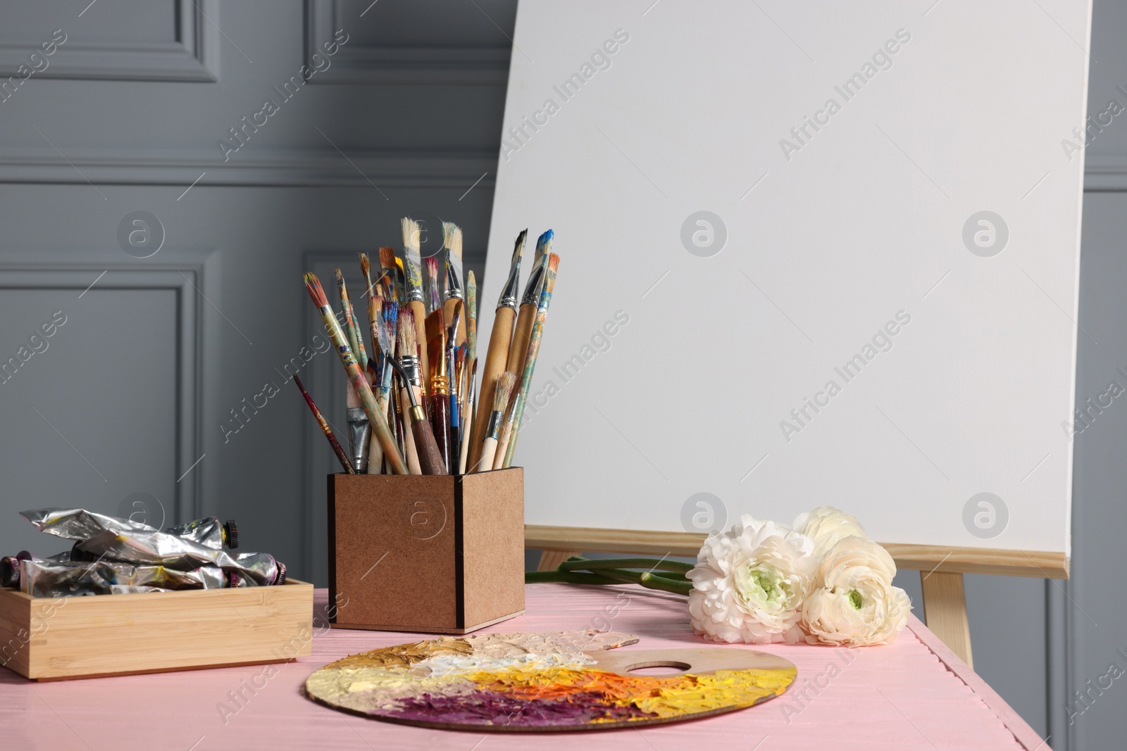 Photo of Easel with blank canvas and different art supplies on wooden table near grey wall