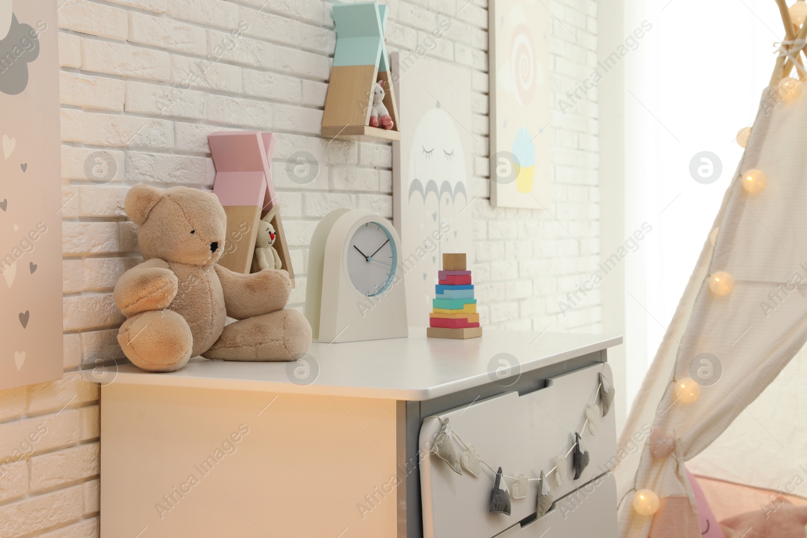 Photo of Toys and clock on chest of drawers near white brick wall in playroom. Interior design