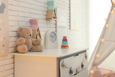 Photo of Toys and clock on chest of drawers near white brick wall in playroom. Interior design