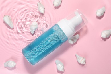 Photo of Bottle of facial cleanser and seashells in water against pink background, flat lay