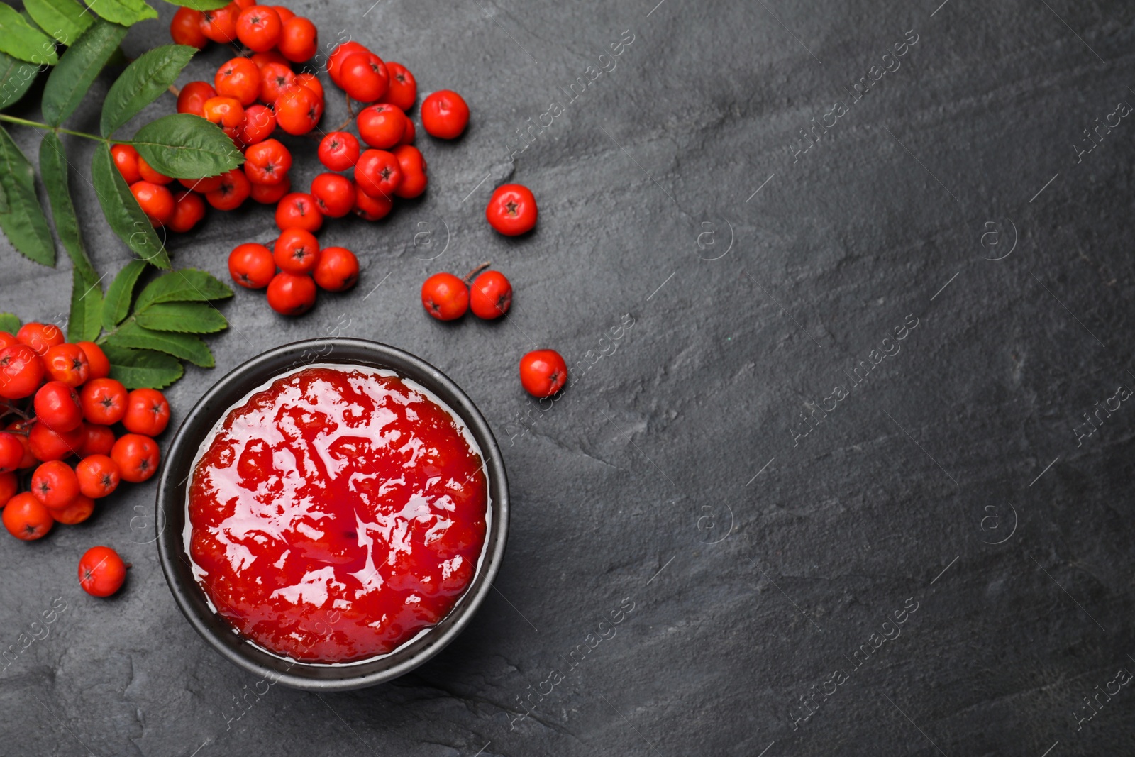 Photo of Delicious rowan jam in bowl and berries on dark table, flat lay. Space for text