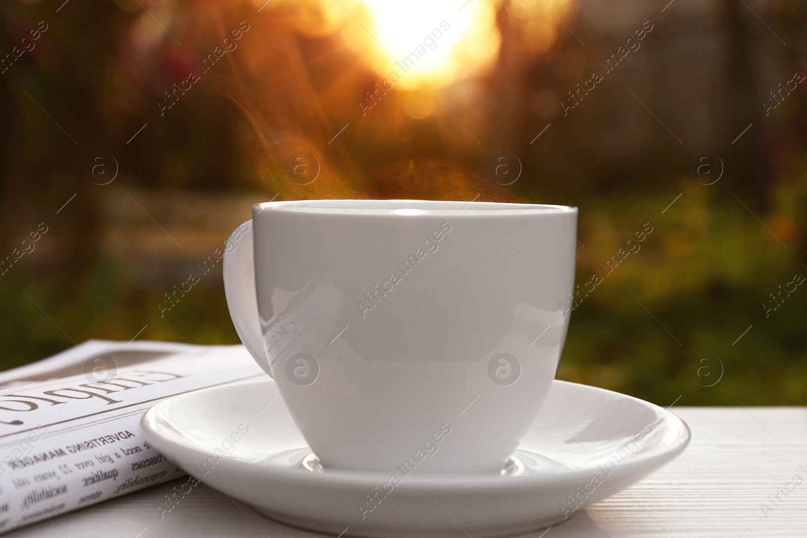 Photo of White cup with coffee and newspaper on wooden table in morning outdoors, closeup