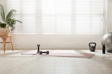 Photo of Exercise mat, dumbbells, kettlebell, fitness ball and bottle near window in spacious room