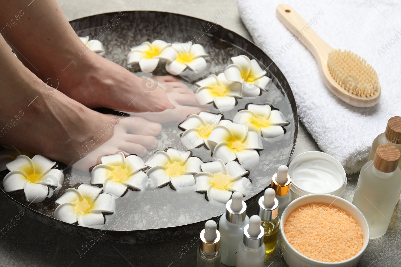 Photo of Woman soaking her feet in bowl with water and flowers on floor, closeup. Spa treatment