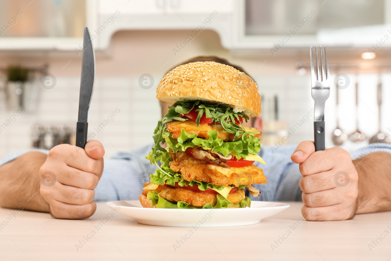 Photo of Young hungry man with cutlery eating huge burger at table