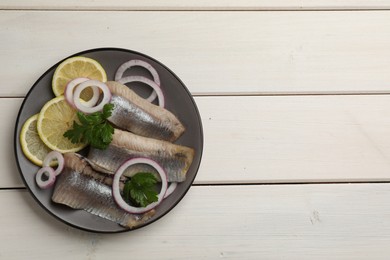 Photo of Delicious salted herring fillets served with lemon, onion rings and parsley on white wooden table, top view. Space for text