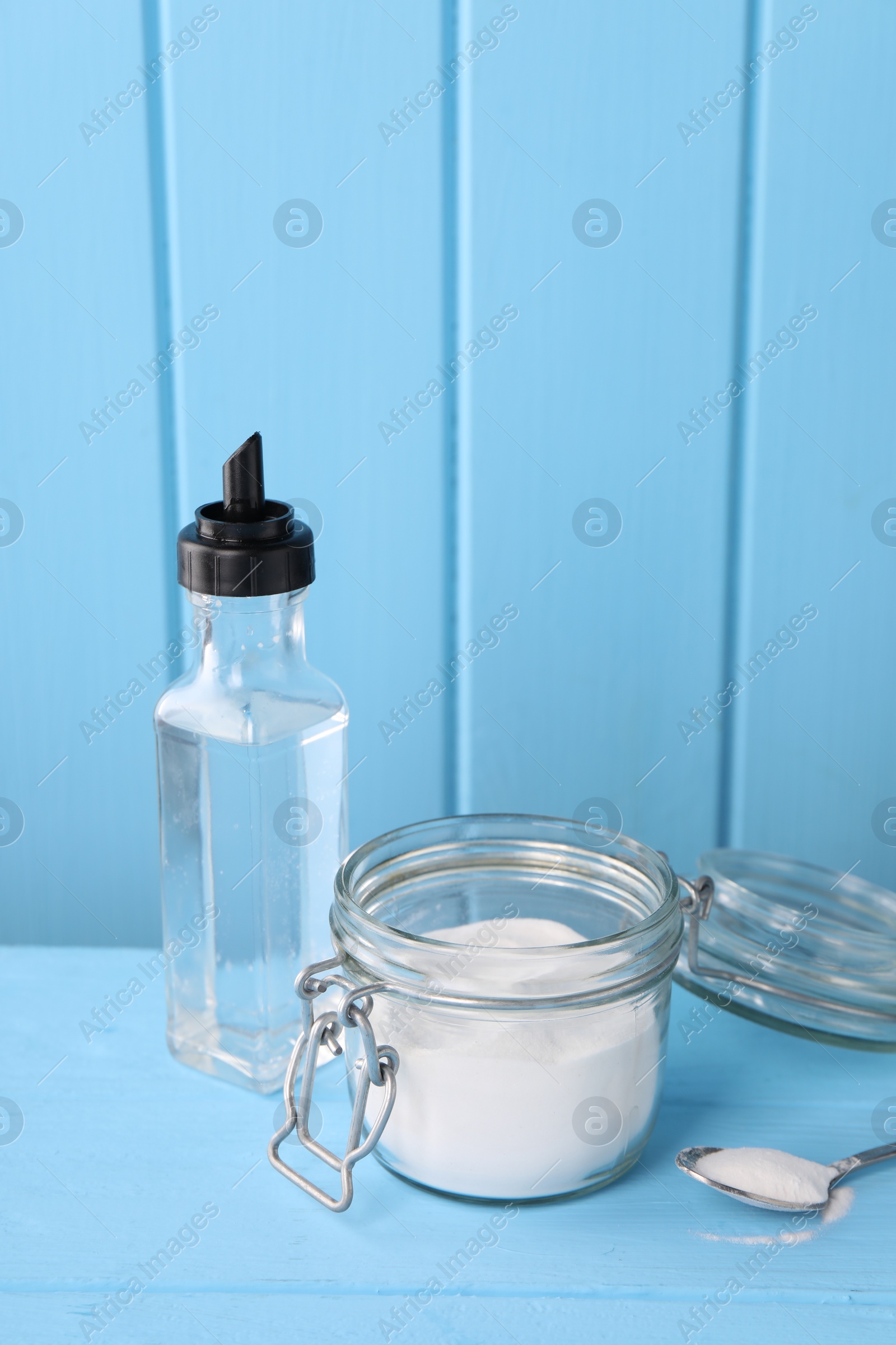 Photo of Natural cleaning products. Vinegar in bottle, baking soda and spoon on light blue wooden table