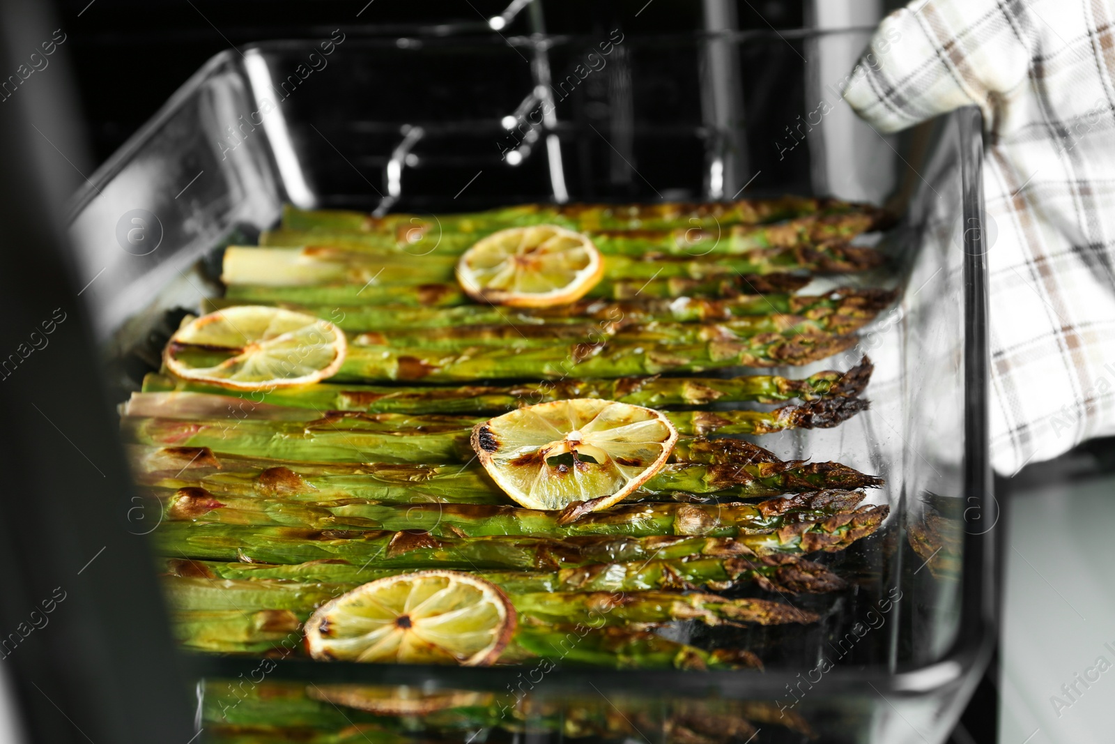 Photo of Person taking glass baking dish with cooked asparagus and lemon slices from oven, closeup