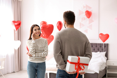 Photo of Young man presenting gift to his girlfriend in bedroom decorated with heart shaped balloons. Valentine's day celebration