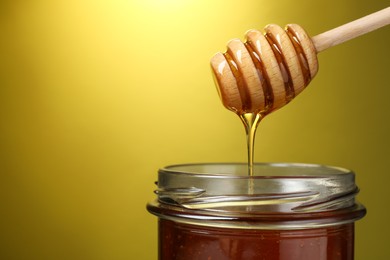 Photo of Pouring tasty honey from dipper into glass jar on golden background, closeup. Space for text
