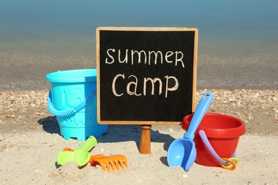 Photo of Small chalkboard with text SUMMER CAMP and beach toys on sand near river