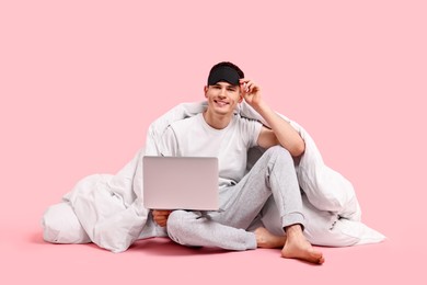 Photo of Happy man in pyjama with sleep mask, blanket and laptop on pink background