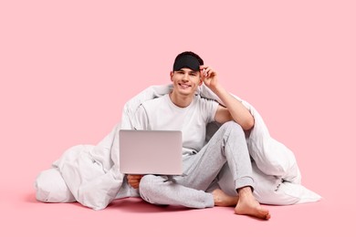 Happy man in pyjama with sleep mask, blanket and laptop on pink background