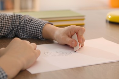 Photo of Boy erasing mistake in his notebook at wooden desk indoors, closeup
