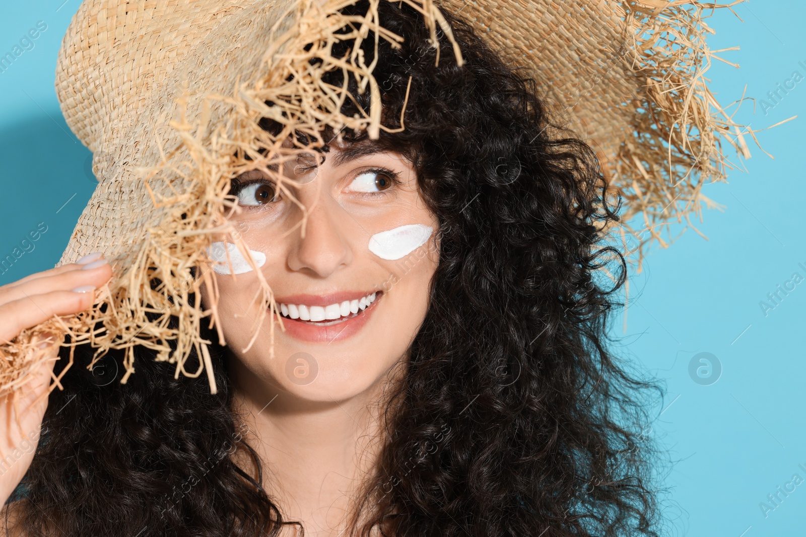Photo of Beautiful young woman in straw hat with sun protection cream on her face against light blue background