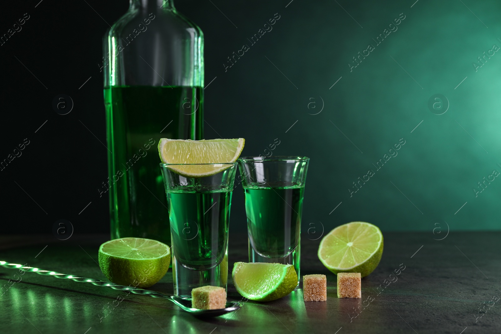 Photo of Absinthe in shot glasses, spoon, brown sugar cubes and lime on gray textured table against green background, space for text. Alcoholic drink