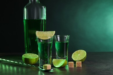 Absinthe in shot glasses, spoon, brown sugar cubes and lime on gray textured table against green background, space for text. Alcoholic drink