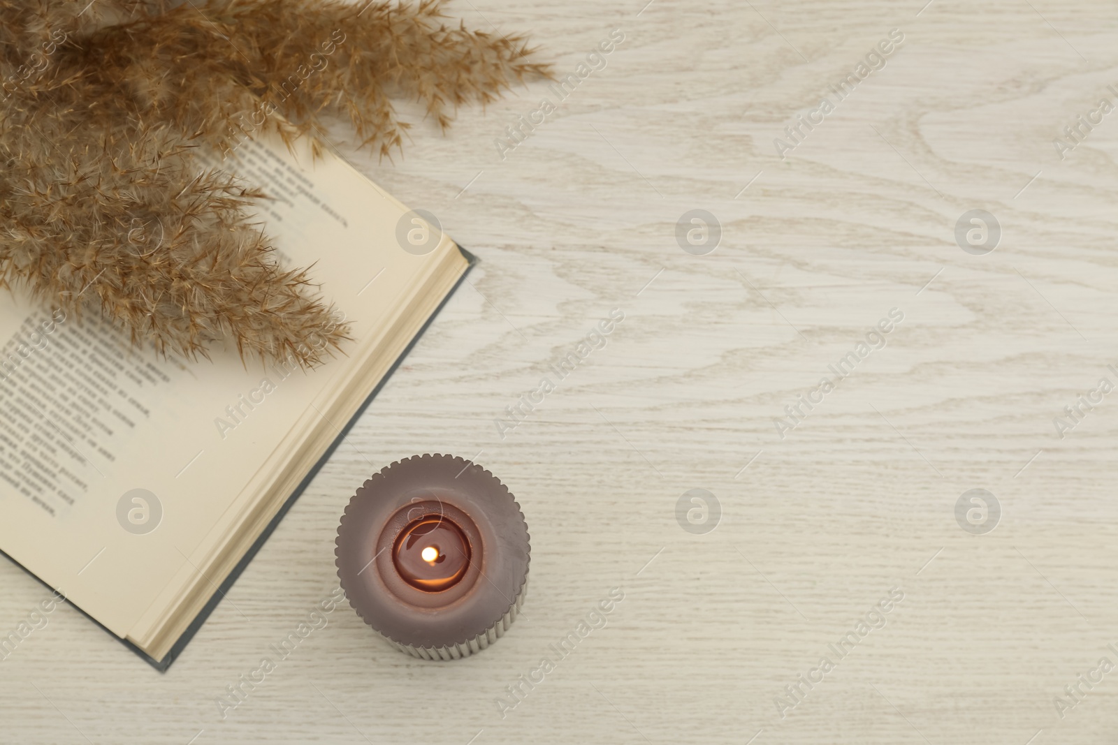 Photo of Burning candle and open book on white wooden table, flat lay. Space for text