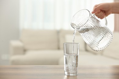 Woman pouring water from jug into glass at table indoors, closeup. Space for text