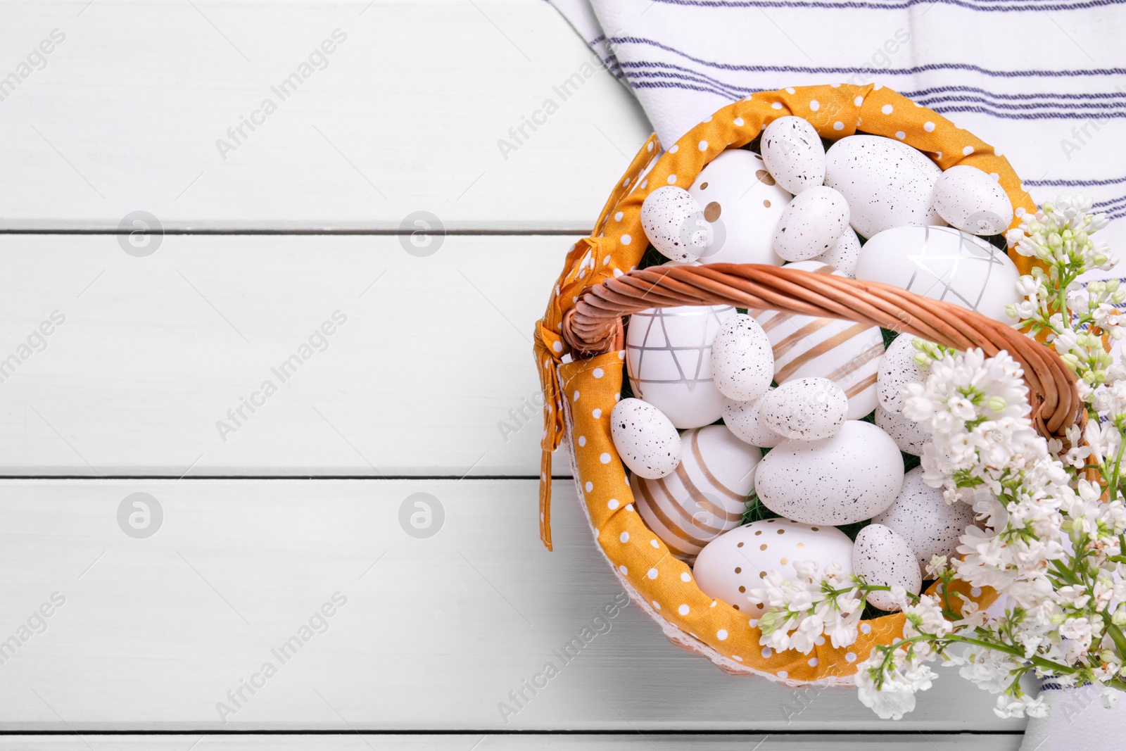 Photo of Easter eggs in basket and lilac flowers on white wooden table, flat lay. Space for text