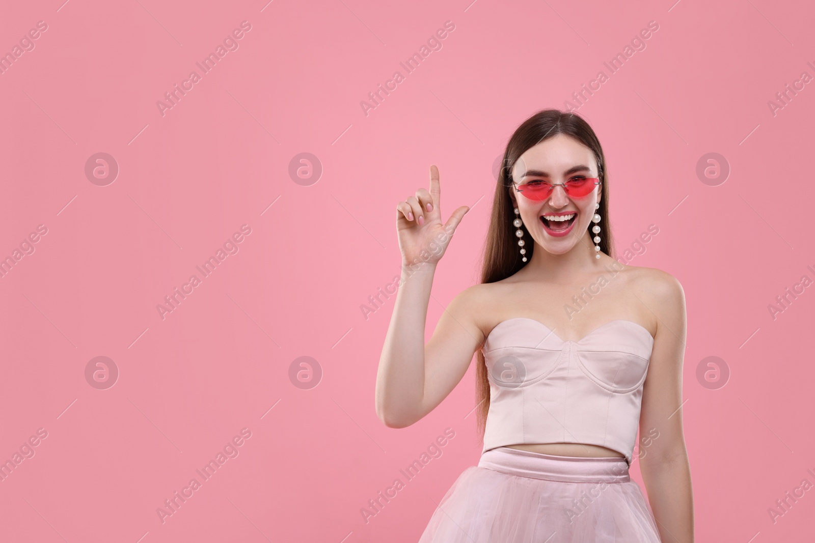 Photo of Pink look. Beautiful woman in clothes and bright sunglasses pointing at something on color background, space for text