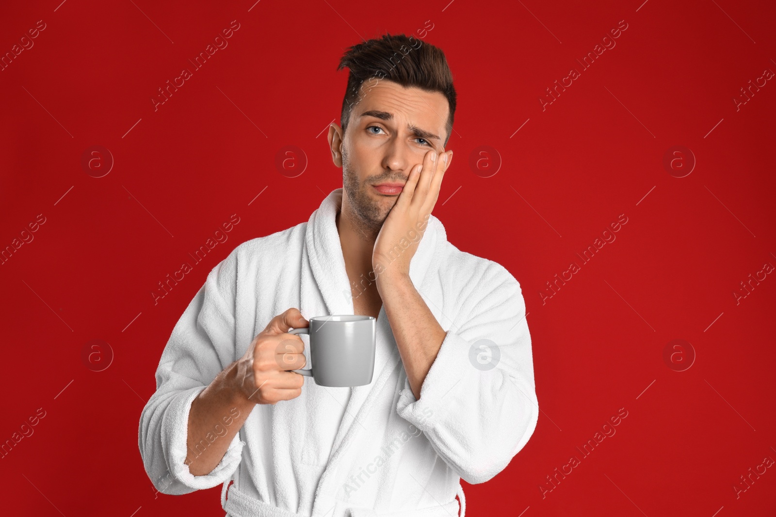 Photo of Sleepy young man in bathrobe  with cup of coffee on red background