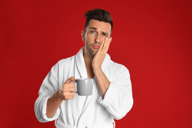 Sleepy young man in bathrobe  with cup of coffee on red background