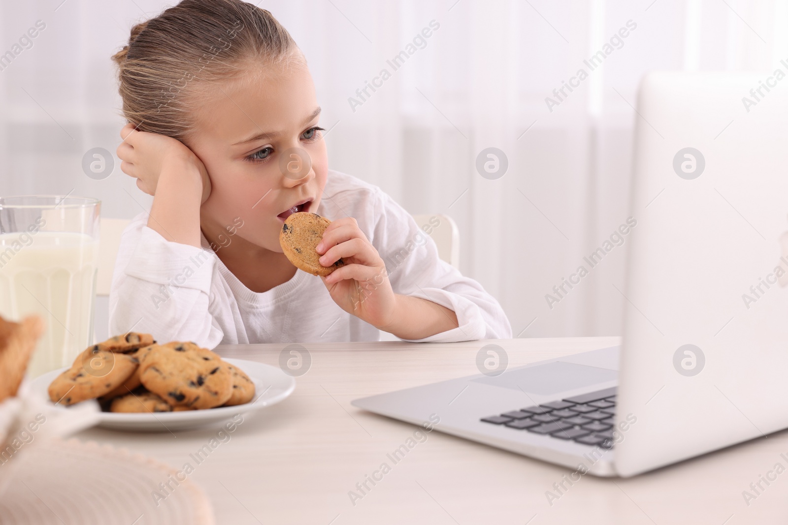 Photo of Little girl using laptop while having breakfast at table indoors. Internet addiction