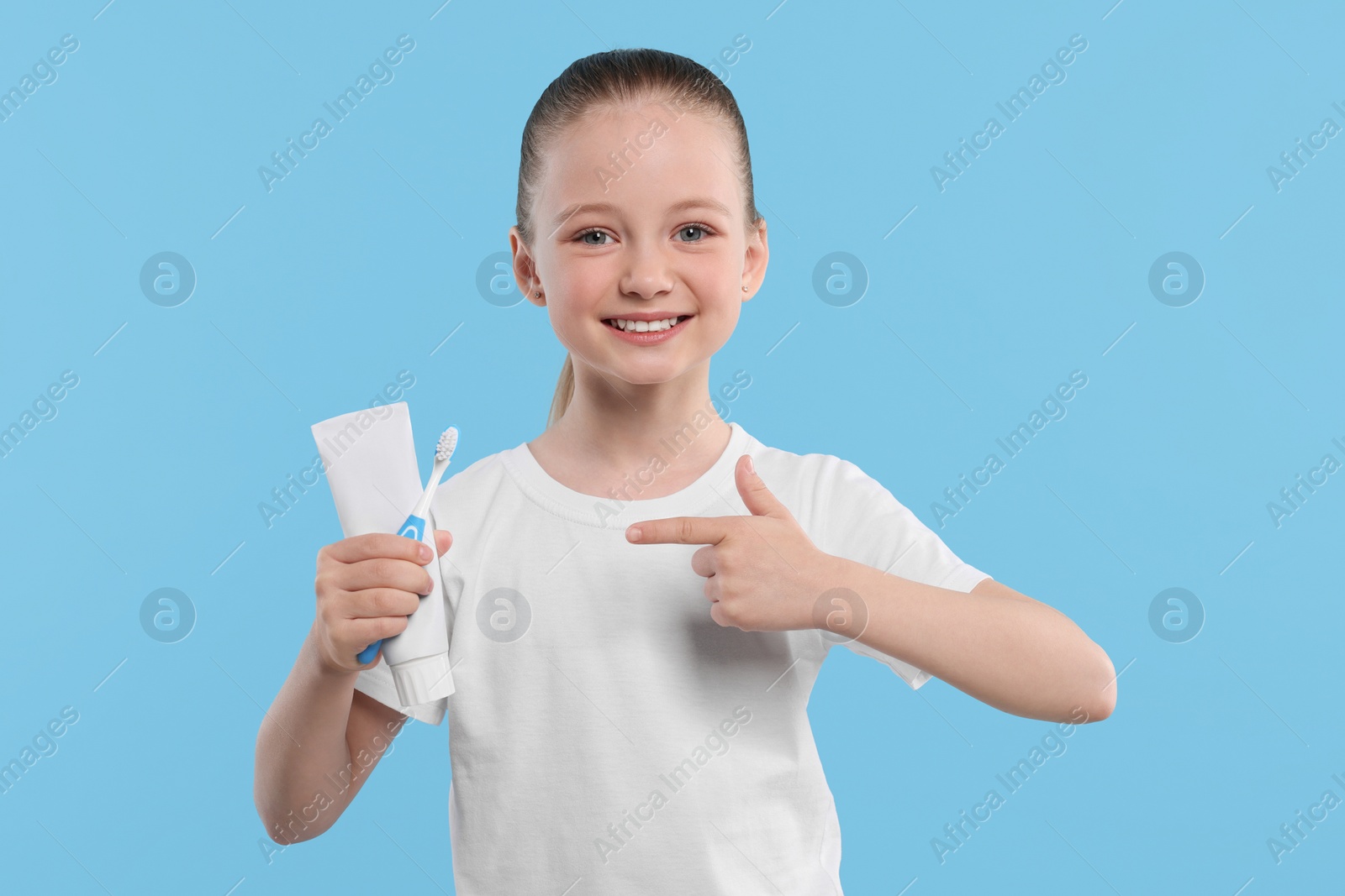 Photo of Happy girl holding toothbrush and tube of toothpaste on light blue background