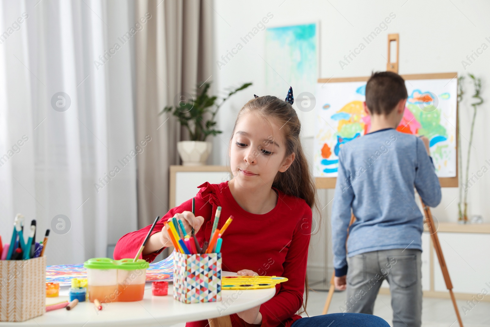 Photo of Girl painting at table and boy near easel indoors. Creative children