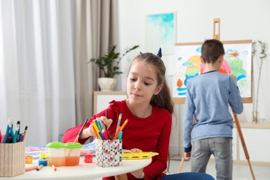 Girl painting at table and boy near easel indoors. Creative children