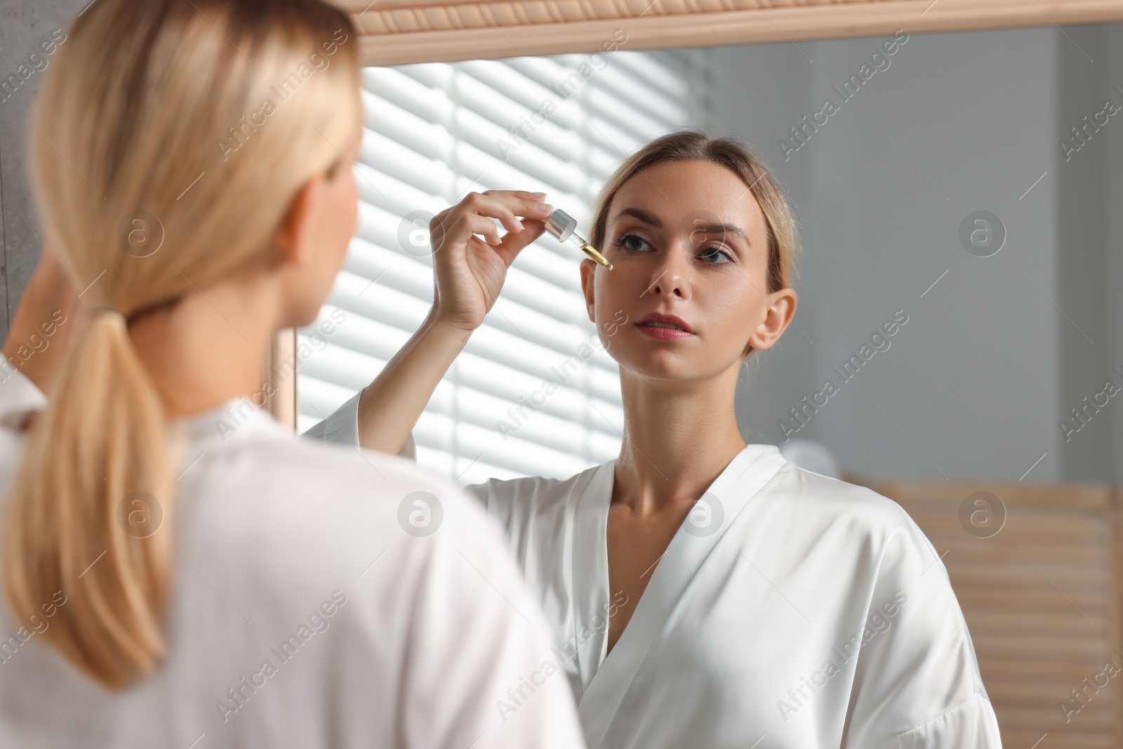 Photo of Beautiful woman applying cosmetic serum onto her face near mirror in bathroom