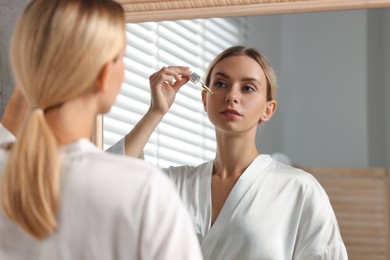 Beautiful woman applying cosmetic serum onto her face near mirror in bathroom