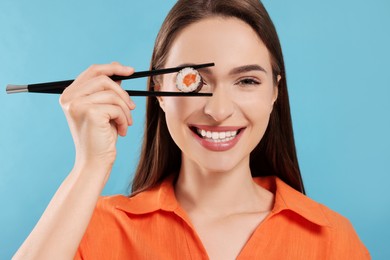 Photo of Funny young woman holding sushi roll with chopsticks on light blue background