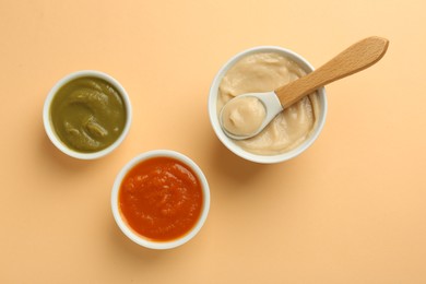 Photo of Bowls with healthy baby food and spoon on pale orange background, flat lay